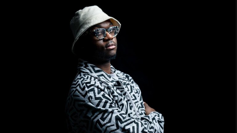 Sultan Stevenson stands against a dark background with his arms crossed, looking toward the camera. He wears a light-colored bucket hat, patterned jacket, and glasses.