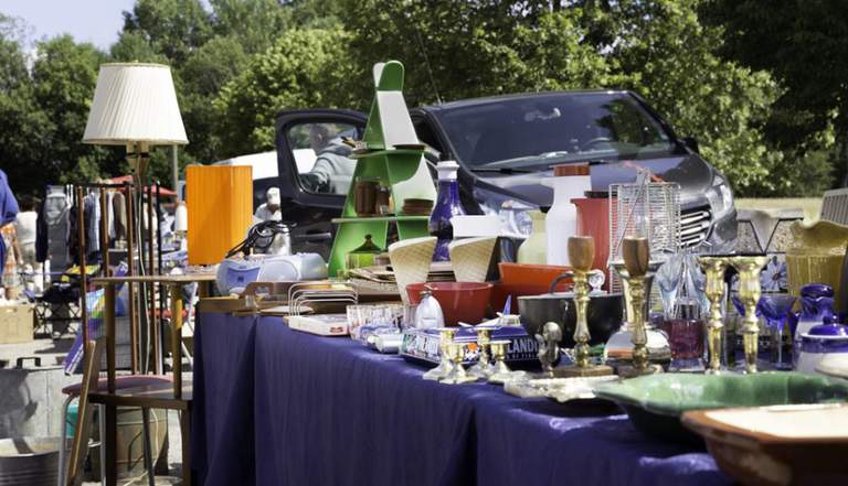 A market stand with silverware, candle holders and more. Green trees and a car can be seenin the bakground.