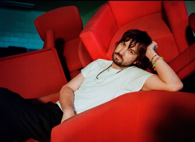 A man in a white t-shirt and colorful bracelets, posing in a sea of red furniture.