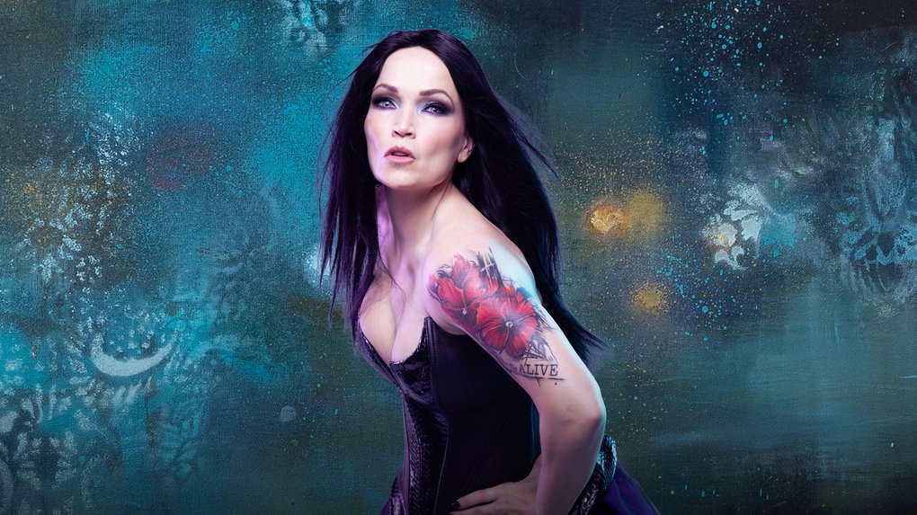 A woman with long black hair poses in front of a blue-green, abstract background. She has a tattoo on her upper arm and is wearing a black dress.