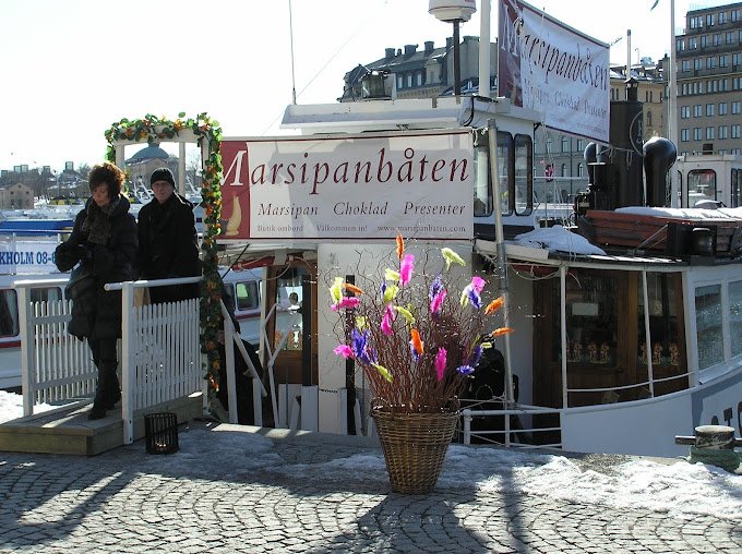 The marzipan boat moored at Nybrokajen in Stockholm with two visitors in thick jackets walking off the gangway. In front stands an Easter egg with feathers. In front of the boat on the quay is a large Easter boquet of branches and feathers.