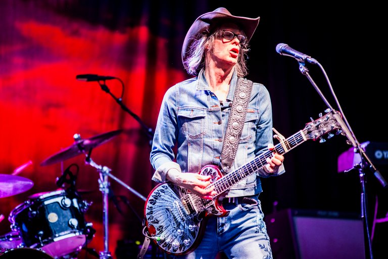 A man in a denim jacket and cowboy hat playing electric guitar and singing on a stage with an amplifier, a keyboard, and a drumset visible behind him.