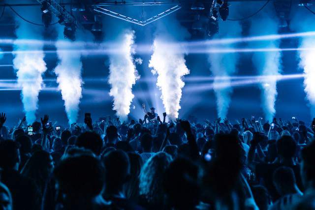 A concert venue with a lot of public and smoke coming up from the stage.