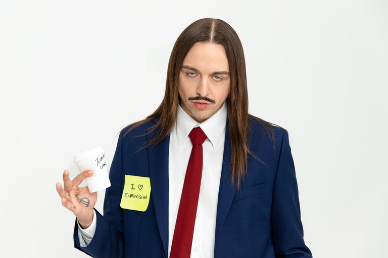 A long-haired man in an ill-fitting suit holding a paper cup.