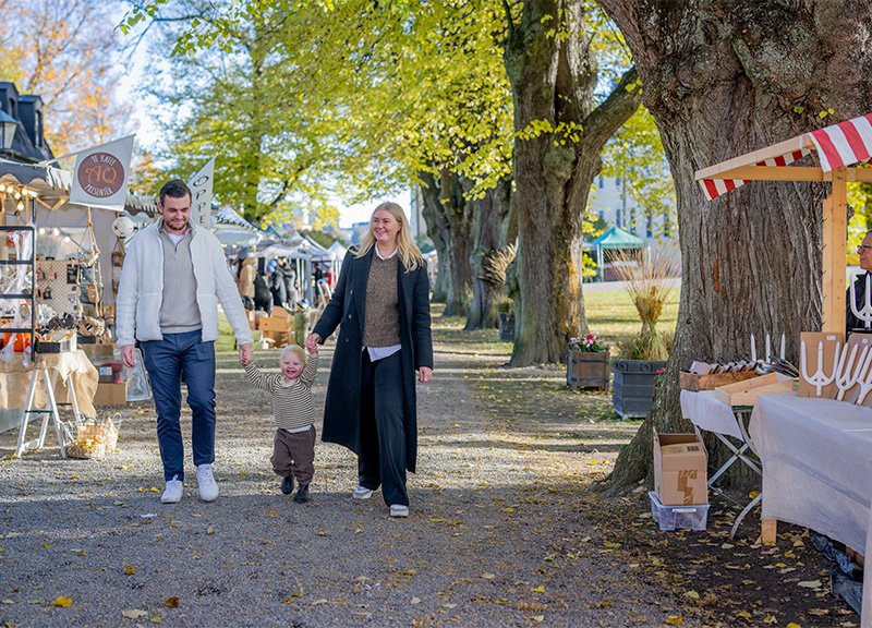 A man and a woman are walking in a park holding hands with a small child. They pass market stalls and trees with autumn-colored leaves.