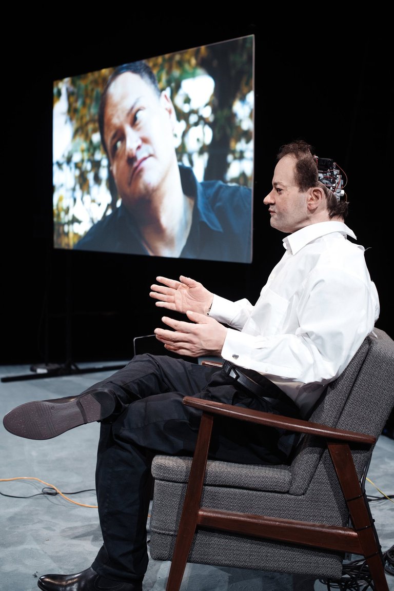 A humanoid robot sitting on a stage. We can see wires in his open skull, and in the background we can see the a picture of author Thomas Melle, on whom the robot is modeled.