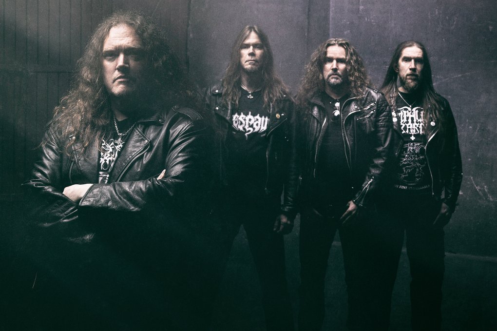 Four long-haired men in leather jackets standing in a dark, slightly smoke-filled room.