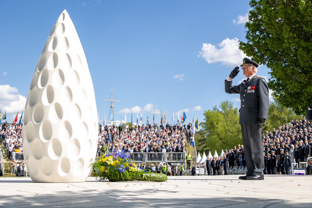 HRM Carl Gustaf XVI of Sweden salutes in front of a white monument during a ceremony with a large audience in the background. A wreath with blue and yellow flowers lies at the base of the monument. The image conveys solemnity, remembrance, and a national ceremony.