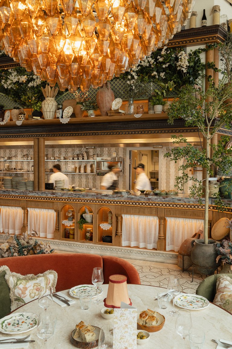 Restaurant interior with a set table, open kitchen counter, lush greenery, and a large glass chandelier in warm light.