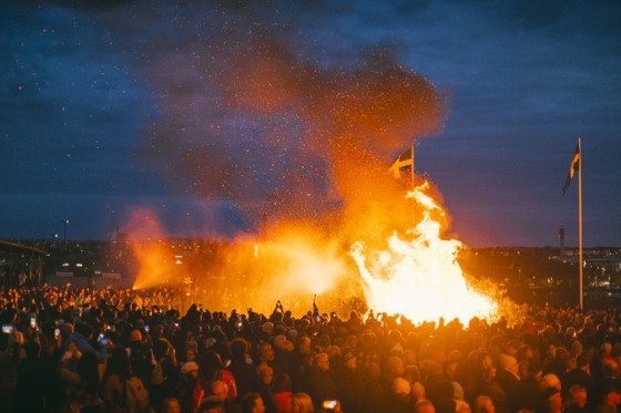 A large crowd gathers around a traditional bonfire during Walpurgis Night celebrations in Stockholm, with Swedish flags visible against the evening sky.