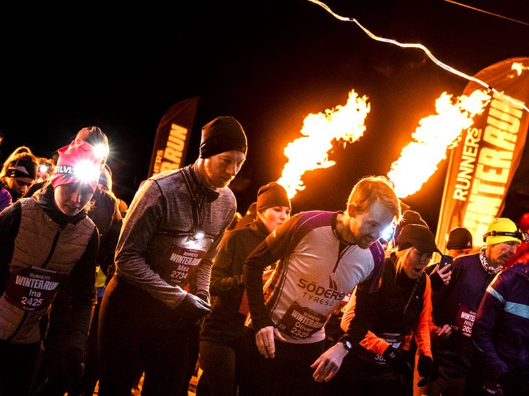 A pack of runners at night, lit by torches and fire performers. Flames and bright lights create a festive winter atmosphere during Winter Run.